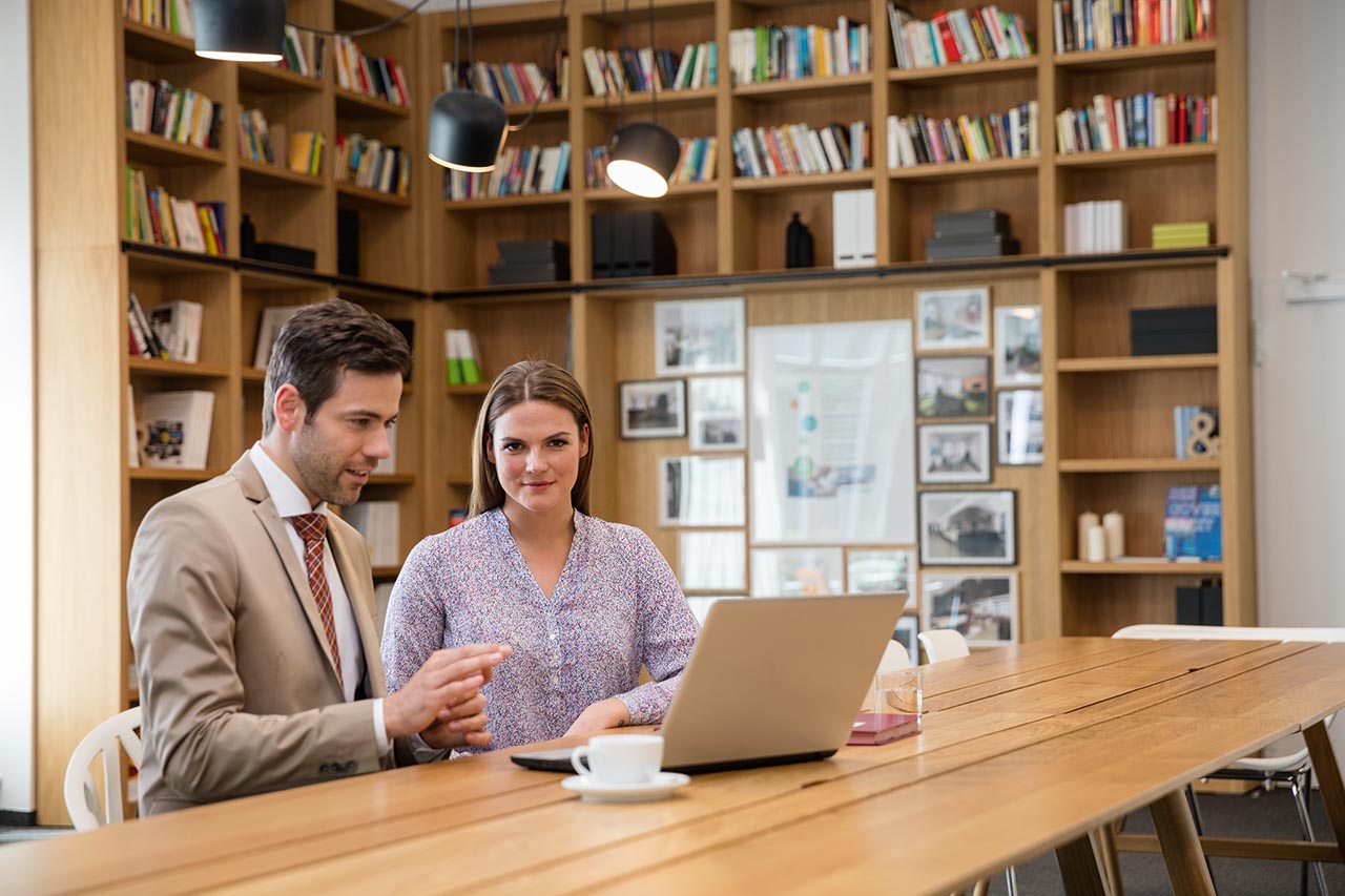 On this picture you can two people sitting in an office in front of a laptop. 