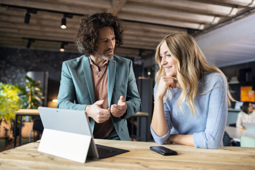 On this picture you can see two persons discussing while standing at a table with a tablet on it.